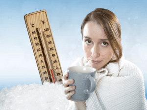 Thermometer stuck in snow behind cold woman covered in blanket and holding a mug