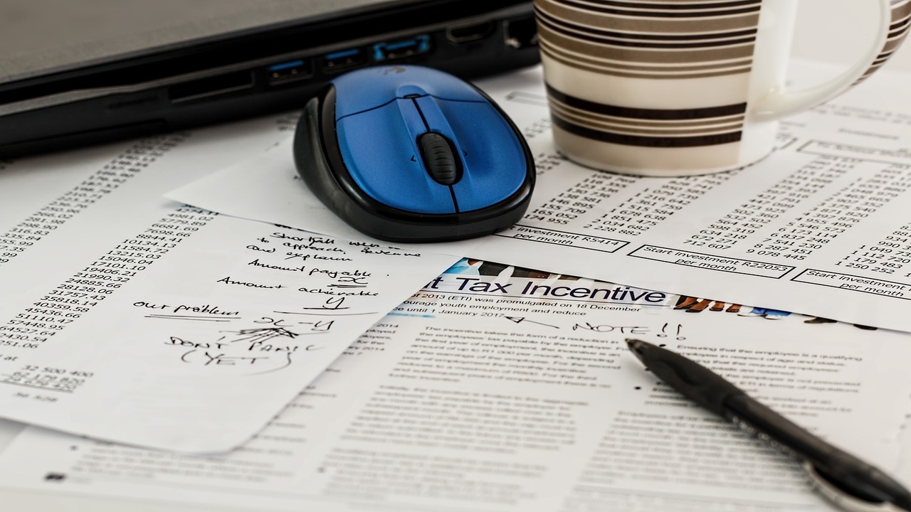 A stock photo showcasing multiple tax forms, a pen, a coffee cup, and a computer mouse.