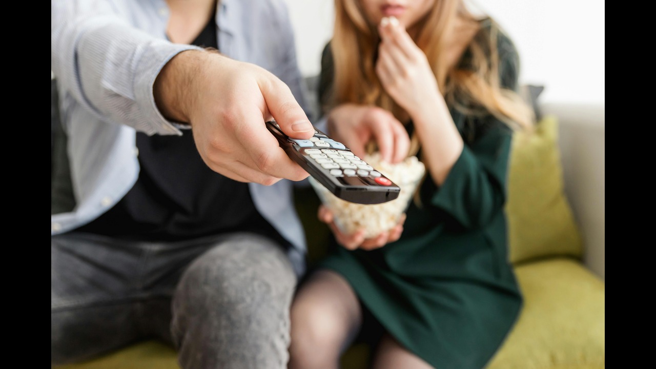 A stock photo showing two people, the person o the left is using a TV remote while the person on the right is eating popcorn.