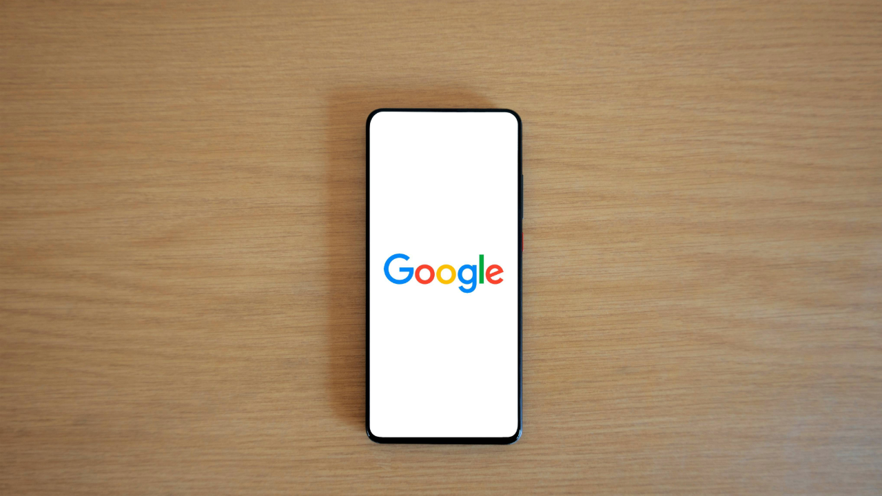 A smartphone showing the Google Logo with a white background. The phone is sitting atop a wooden table.