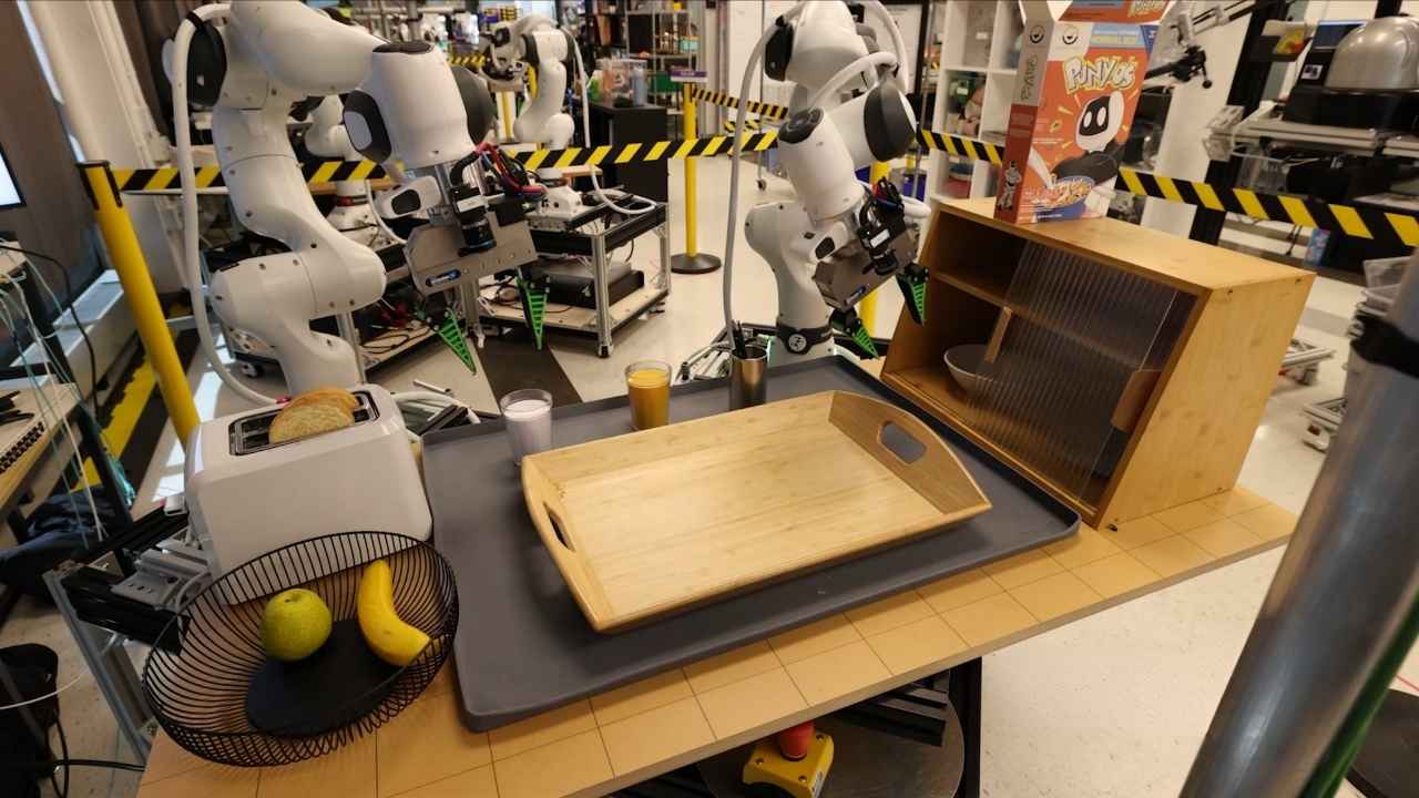 Robotic arms work around a kitchen-style test area, showing how much setup and supervision physical robot training can require.